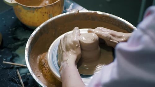 Close-up of potter's hands covered with clay making beautiful vase on throwing wheel in pottery work
