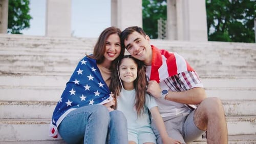 Portrait of A Young Family with a Child Girl is Sitting on the Steps and Wrapped in the Flag of the