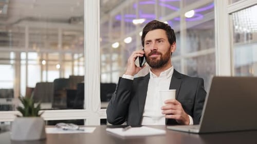 Businessman Talking on Phone and Drinking Coffee at Desk in Modern Office