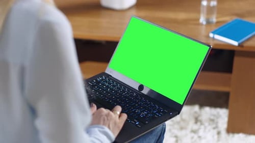 Over the Shoulder Shot of a Woman Working on a Laptop with Green Mock-up Screen on it. She Holds La