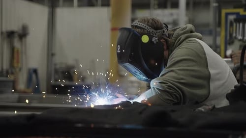 Worker wearing face shield welding metal, bright sparks flying in workshop. Slow motion