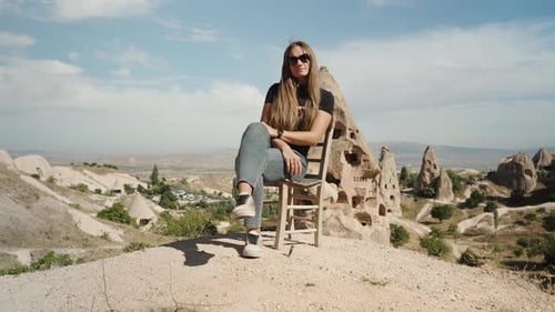 Una mujer joven se sienta en una roca alta sobre una silla de madera, con el fondo de una ciudad antigua.
