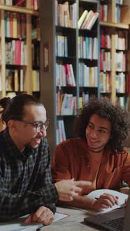 Two Male Students Using Laptop and Discussing Homework in Library