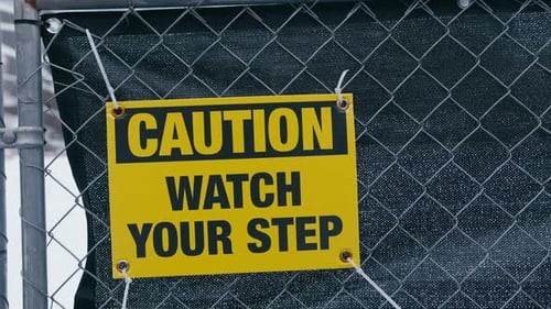A Yellow Caution Sign Reading Caution Watch Your Step Mounted on a Chainlink Fence and Secured with