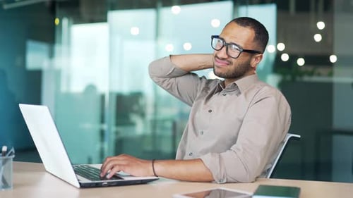 Young adult man suffering from neck pain while sitting at workplace at desk in modern office. A mixe