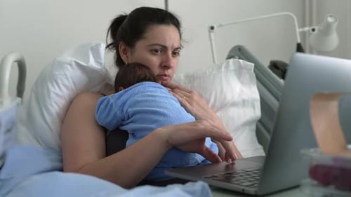 New mother multitasking in hospital bed with newborn baby, working on laptop, combining parenting