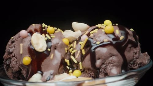 Ein Glas mit Schokoladeneiskugeln und Zuckerstreuseln dreht sich auf schwarzem Hintergrund, Nahaufnahme