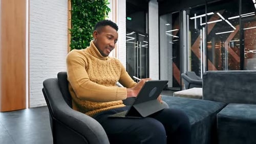 Black man working using a tablet while sitting on a sofa in an office. Slow motion