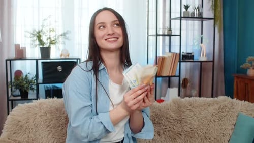 Young Woman Dancing Joyfully on Sofa Holding Cash Euro Bills Celebrating Lottery Winning Success