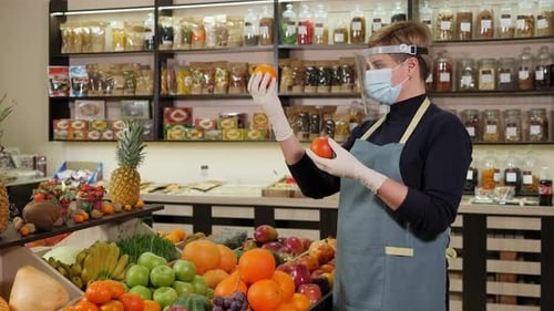 A Saleswoman in a Mask with a Shield and Gloves Holds an Orange in a Store