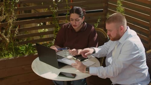 Coworkers Reviewing Documents at Table with Laptop in Outdoor Cafe