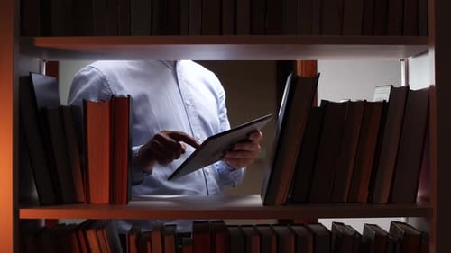 Man Using Tablet Between Bookshelves in Library at Night