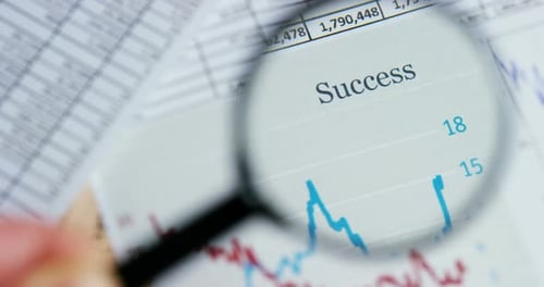 A hand with a magnifying glass passes over a financial newspaper and financial bag with a business