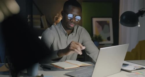 African American Businessman Using Laptop and Writing Information in Notebook in Office in Evening