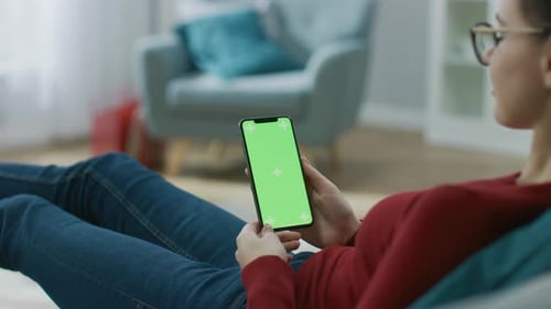 Young Woman at Home Uses Green Mock-up Screen Smartphone. She's Sitting On a Couch in His Cozy Livi
