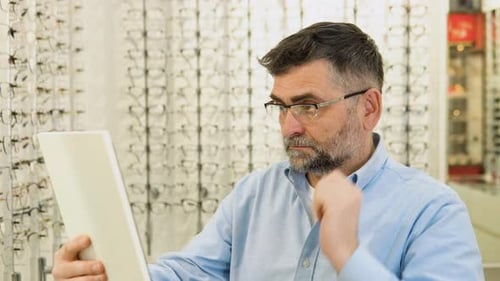 Hombre mayor eligiendo gafas nuevas en una tienda de óptica mirándose al espejo