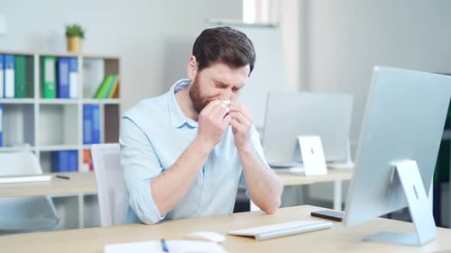 Bearded Man Blowing Nose at Desk in Office