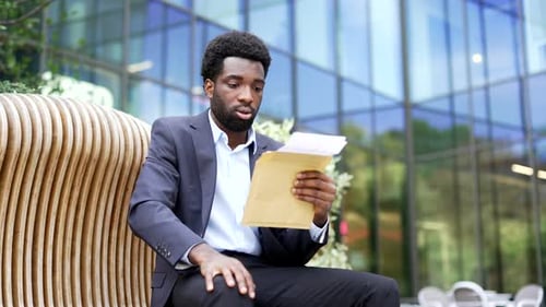 Worried sad african american businessman reading letter with bad news sitting on bench on street