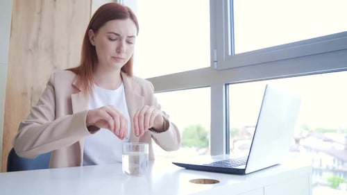 young caucasian woman drinking painkillers with water at workplace. Businesswoman sitting at office