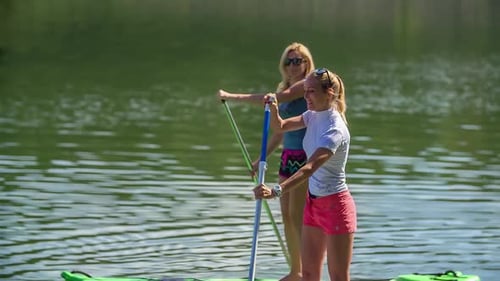 Two Female Friends Standing Up On Paddle Boards Row By. Follow Shot, Slow Motion