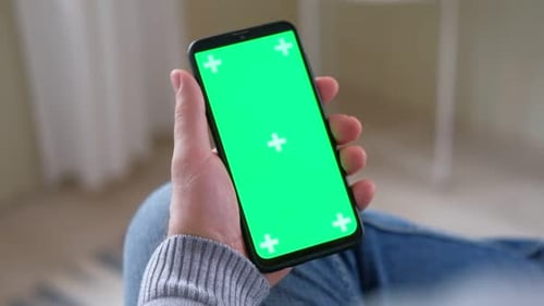 Young Man Sitting at Home Holding Smartphone Green Mockup Screen in Hand Male Person Using Chroma
