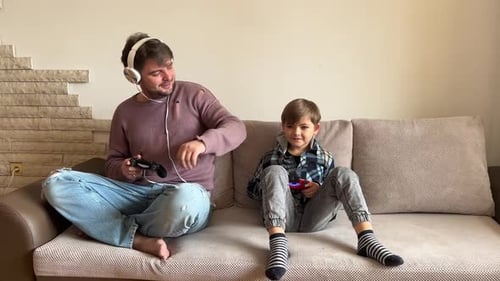 Father And Son Sitting On Sofa In Lounge Playing Video Game