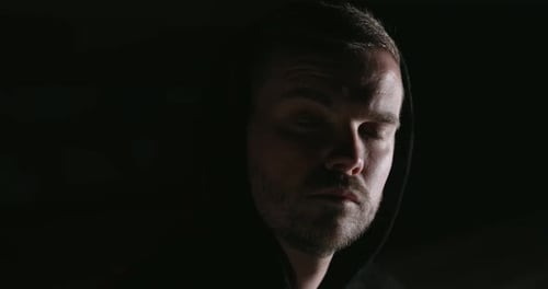 A man in the dark looks into the camera. his face is half-lit. he shows various emotions