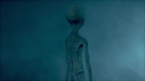 Scary gray alien standing and blinking at the dark foggy background of a moon-like planet