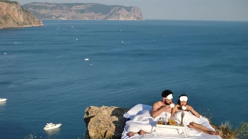 Desayuno romántico de un hombre y una mujer en una cama blanca en un acantilado con vistas al mar y los yates