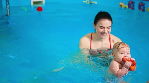 Positive woman is moving her baby in the swimming pool. Little infant boy is chewing a toy.