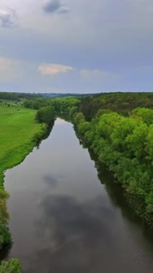 Narrow river with meadow on one side and forest on another. Cloudy sky reflecting in the water.