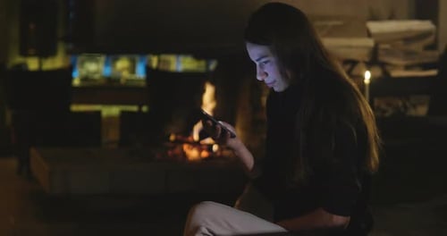 Young Woman Using Phone by Fireplace at Night