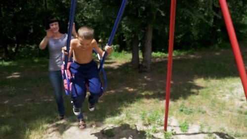 Mother Swinging Her Happy Child Boy on a Street Swing at Playground in Slow Motion