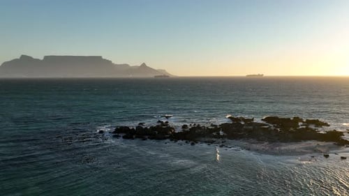 Drone flight above rocky rugged reef with distant sunset view of Table mountain