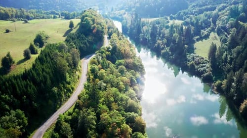 Winding country road along bright blue river in which the bright sun is reflected surrounded by gree