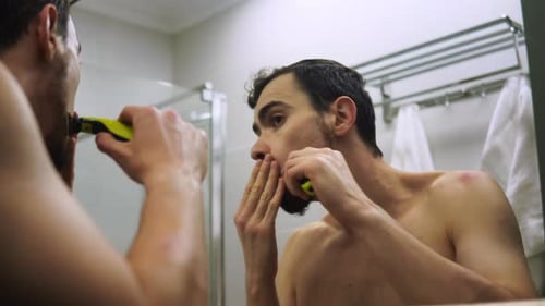 Caucasian Man is Shaving Beard By Razor Standing Shirtless in Bathroom in Front of Mirror