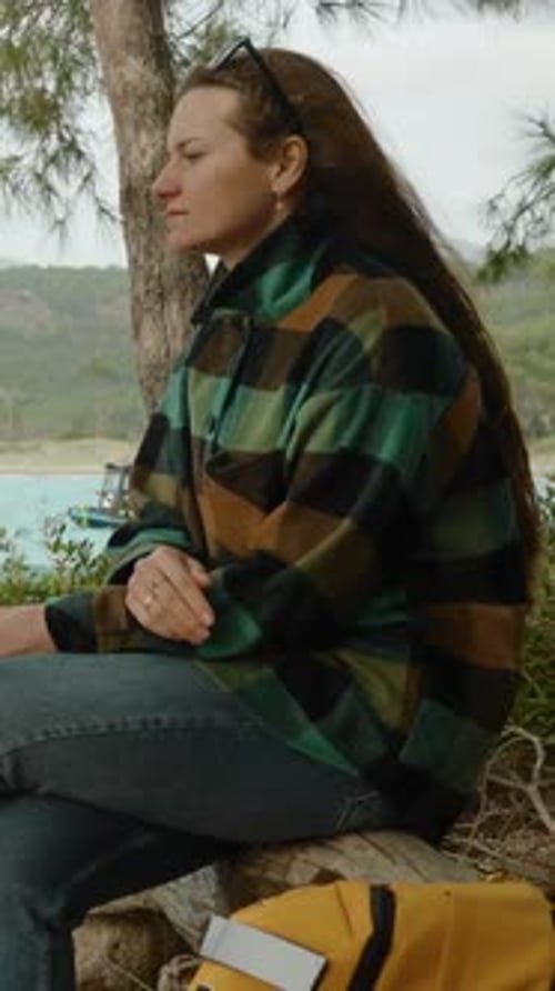 Vertical video. A female traveler sits on a fallen tree in the forest by the sea, lost in thought.