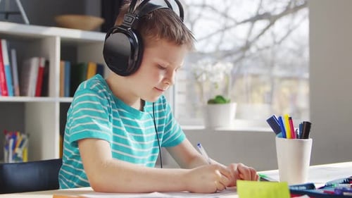 Young Boy Writes at Desk Wearing Headphones