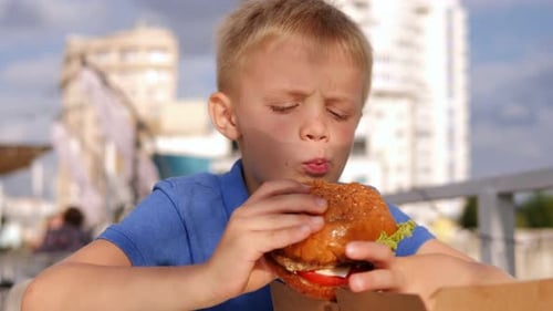 A Little Boy Eats a Big Burger in an Outdoor Restaurant in Summer