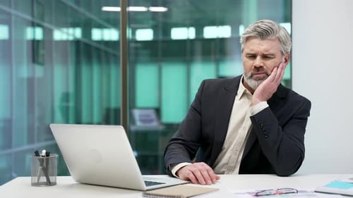 Mature businessman suffering from toothache while sitting at workplace at desk in business office.