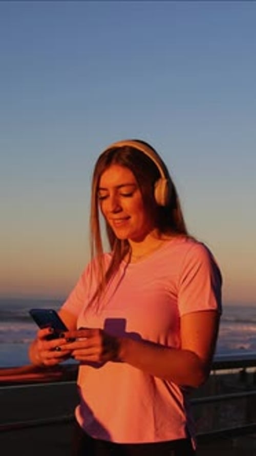 vertical Young woman using smartphone and listening music with headphones at sunset