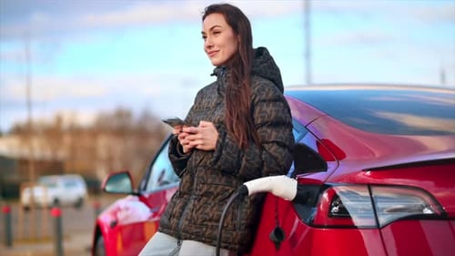 Ansicht einer Frau, die ein Smartphone in der Nähe ihres aufladenden roten Elektroautos benutzt. Zeitlupe