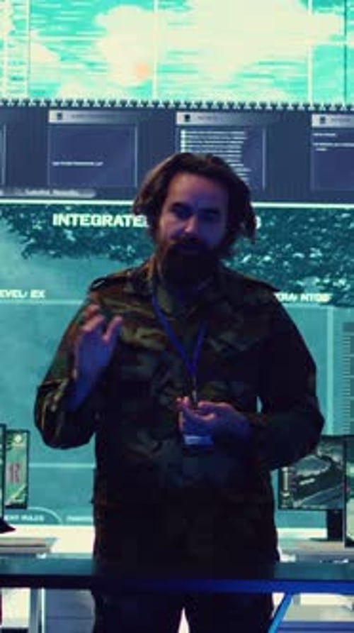 Man in Camouflage Jacket Stands in Front of Screens