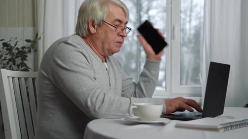 Seriöser älterer Mann mit Brille, sprechendes Handy, sitzt am Schreibtisch zu Hause am Arbeitsplatz mit