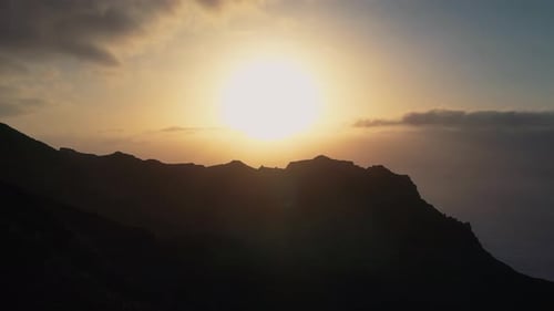 Aerial View Past Silhouette Of Adult Standing On Mountainside With Golden Yellow Sunset In The Backg