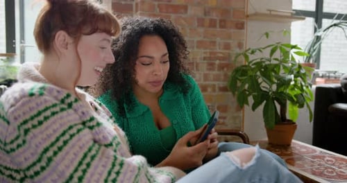 Two Young adult Female Friends looking at a Smartphone Together in a shared loft apartment on Sofa