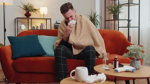 Kranker kranker Kerl, der an Erkältung oder Allergie leidet, auf dem Sofa zu Hause liegend, niest, wischt Rotz in die Serviette