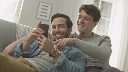 Cute Male Gay Couple Spend Time at Home. They are Lying Down on a Sofa and Use a Smartphone. They B