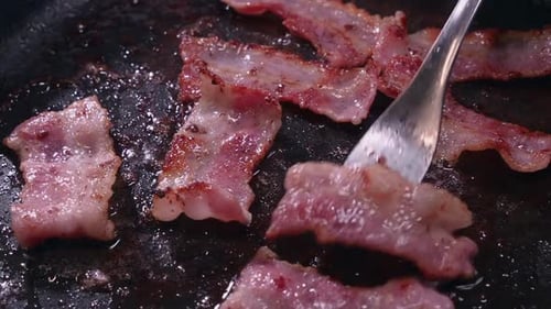 Tasty bacon sizzles in hot frying pan as fork flips strips, closeup