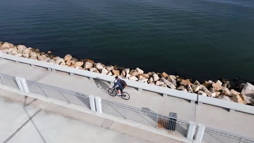 A Man with a Backpack Rides a Bicycle Along a Seafront Promenade Seen From a Flying Drone